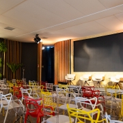 O ambiente mostra um audit&oacute;rio preparado para palestras, com cadeiras coloridas distribu&iacute;das em frente a um palco. O palco possui uma tela grande ao centro, quatro cadeiras altas e decora&ccedil;&atilde;o com pain&eacute;is de madeira vertical.