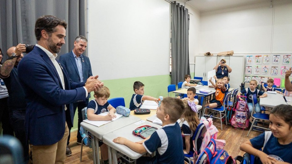 A fotografia registra um momento em uma sala de aula de uma escola prim&aacute;ria, com a presen&ccedil;a do Governador do Rio Grande do Sul, Eduardo Leite, e outros adultos interagindo com crian&ccedil;as.

Elementos Principais
Primeiro Plano (Esquerda): O governador Eduardo Leite, um homem branco de cabelos escuros e barba curta, veste um blazer azul-marinho sobre uma camisa branca e cal&ccedil;as c&aacute;qui. Ele est&aacute; de perfil, sorrindo e batendo palmas em dire&ccedil;&atilde;o aos alunos.

O Grupo de Alunos: Cerca de dez crian&ccedil;as, com idades estimadas entre 6 e 8 anos, est&atilde;o sentadas em duplas ou pequenos grupos em suas classes. A maioria veste o uniforme escolar: uma camiseta azul-marinho com detalhes brancos nas mangas.

Intera&ccedil;&atilde;o: No centro, um menino est&aacute; sentado &agrave; mesa escrevendo, enquanto outro, logo &agrave; frente, olha para cima em dire&ccedil;&atilde;o aos adultos. Ao fundo, uma menina com uma camiseta laranja vibrante levanta os bra&ccedil;os com entusiasmo, comemorando ou respondendo a algo.

O Ambiente (Sala de Aula)
Mobili&aacute;rio: As classes s&atilde;o cinza-claras com estruturas met&aacute;licas, acompanhadas de cadeiras azuis. Sobre as mesas, veem-se estojos, cadernos e garrafas de &aacute;gua. Mochilas coloridas est&atilde;o penduradas nas cadeiras ou no ch&atilde;o.

Decora&ccedil;&atilde;o e Estrutura: As paredes s&atilde;o brancas com um barrado verde-claro na parte inferior. Ao fundo, h&aacute; um arm&aacute;rio branco e, na parede lateral, diversos desenhos e atividades escolares coloridas est&atilde;o pendurados em um varal did&aacute;tico. Cortinas cinzas cobrem as janelas ao fundo e &agrave; esquerda.

Presen&ccedil;a de M&iacute;dia: &Agrave; esquerda, atr&aacute;s do governador, v&ecirc;-se parte do bra&ccedil;o de um fot&oacute;grafo segurando uma c&acirc;mera profissional, indicando que se trata de uma visita oficial ou evento p&uacute;blico.

Atmosfera
A ilumina&ccedil;&atilde;o &eacute; clara e uniforme, sugerindo luz artificial de teto. O clima geral &eacute; de alegria e engajamento, com express&otilde;es sorridentes tanto dos adultos quanto das crian&ccedil;as.