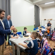 A fotografia registra um momento em uma sala de aula de uma escola prim&aacute;ria, com a presen&ccedil;a do Governador do Rio Grande do Sul, Eduardo Leite, e outros adultos interagindo com crian&ccedil;as.

Elementos Principais
Primeiro Plano (Esquerda): O governador Eduardo Leite, um homem branco de cabelos escuros e barba curta, veste um blazer azul-marinho sobre uma camisa branca e cal&ccedil;as c&aacute;qui. Ele est&aacute; de perfil, sorrindo e batendo palmas em dire&ccedil;&atilde;o aos alunos.

O Grupo de Alunos: Cerca de dez crian&ccedil;as, com idades estimadas entre 6 e 8 anos, est&atilde;o sentadas em duplas ou pequenos grupos em suas classes. A maioria veste o uniforme escolar: uma camiseta azul-marinho com detalhes brancos nas mangas.

Intera&ccedil;&atilde;o: No centro, um menino est&aacute; sentado &agrave; mesa escrevendo, enquanto outro, logo &agrave; frente, olha para cima em dire&ccedil;&atilde;o aos adultos. Ao fundo, uma menina com uma camiseta laranja vibrante levanta os bra&ccedil;os com entusiasmo, comemorando ou respondendo a algo.

O Ambiente (Sala de Aula)
Mobili&aacute;rio: As classes s&atilde;o cinza-claras com estruturas met&aacute;licas, acompanhadas de cadeiras azuis. Sobre as mesas, veem-se estojos, cadernos e garrafas de &aacute;gua. Mochilas coloridas est&atilde;o penduradas nas cadeiras ou no ch&atilde;o.

Decora&ccedil;&atilde;o e Estrutura: As paredes s&atilde;o brancas com um barrado verde-claro na parte inferior. Ao fundo, h&aacute; um arm&aacute;rio branco e, na parede lateral, diversos desenhos e atividades escolares coloridas est&atilde;o pendurados em um varal did&aacute;tico. Cortinas cinzas cobrem as janelas ao fundo e &agrave; esquerda.

Presen&ccedil;a de M&iacute;dia: &Agrave; esquerda, atr&aacute;s do governador, v&ecirc;-se parte do bra&ccedil;o de um fot&oacute;grafo segurando uma c&acirc;mera profissional, indicando que se trata de uma visita oficial ou evento p&uacute;blico.

Atmosfera
A ilumina&ccedil;&atilde;o &eacute; clara e uniforme, sugerindo luz artificial de teto. O clima geral &eacute; de alegria e engajamento, com express&otilde;es sorridentes tanto dos adultos quanto das crian&ccedil;as.