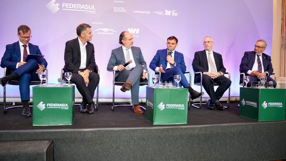 Esta &eacute; uma descri&ccedil;&atilde;o detalhada da imagem para fins de acessibilidade:

A fotografia registra um painel de discuss&atilde;o durante o 11&ordm; Encontro de Embaixadores, realizado pela Federasul.

Composi&ccedil;&atilde;o e Elementos Centrais
O Painel: Seis homens est&atilde;o sentados em cadeiras pretas sobre um palco elevado. Eles est&atilde;o organizados em linha, vestindo trajes formais (ternos e palet&oacute;s em tons de azul e preto).

Intera&ccedil;&atilde;o: O quarto homem da esquerda para a direita (sentado ao centro-direita) est&aacute; falando ao microfone, gesticulando com a m&atilde;o livre. Os demais integrantes do painel o observam ou mant&ecirc;m uma postura de escuta atenta.

Mobili&aacute;rio do Palco: &Agrave; frente dos palestrantes, h&aacute; tr&ecirc;s cubos verdes que servem como mesas de apoio, exibindo o logotipo da FEDERASUL em branco. Sobre esses cubos, encontram-se copos de &aacute;gua e microfones extras.

Plano de Fundo e Identifica&ccedil;&atilde;o
Painel de Fundo: Um grande banner em tons de lil&aacute;s e roxo claro preenche o fundo. No topo, l&ecirc;-se parcialmente a palavra "EMBAIXADORES".

Logotipos Institucionais: O banner exibe diversas marcas de realizadores e apoiadores, incluindo:

FEDERASUL (Realiza&ccedil;&atilde;o)

SOUTH SUMMIT e INVEST RS (Apoiadores)

Governo do Estado do Rio Grande do Sul / Secretaria de Desenvolvimento Econ&ocirc;mico

Logotipos de patrocinadores como AEROMOT e W3.

Ambiente e Ilumina&ccedil;&atilde;o
O palco tem um acabamento frontal em carpete cinza escuro.

A ilumina&ccedil;&atilde;o &eacute; focada nos participantes, t&iacute;pica de eventos de audit&oacute;rio, criando um contraste n&iacute;tido com o fundo decorativo.

O clima &eacute; de um evento corporativo e diplom&aacute;tico formal, focado em discuss&otilde;es estrat&eacute;gicas e desenvolvimento econ&ocirc;mico.