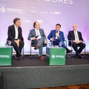 Esta &eacute; uma descri&ccedil;&atilde;o detalhada da imagem para fins de acessibilidade:

A fotografia registra um painel de discuss&atilde;o durante o 11&ordm; Encontro de Embaixadores, realizado pela Federasul.

Composi&ccedil;&atilde;o e Elementos Centrais
O Painel: Seis homens est&atilde;o sentados em cadeiras pretas sobre um palco elevado. Eles est&atilde;o organizados em linha, vestindo trajes formais (ternos e palet&oacute;s em tons de azul e preto).

Intera&ccedil;&atilde;o: O quarto homem da esquerda para a direita (sentado ao centro-direita) est&aacute; falando ao microfone, gesticulando com a m&atilde;o livre. Os demais integrantes do painel o observam ou mant&ecirc;m uma postura de escuta atenta.

Mobili&aacute;rio do Palco: &Agrave; frente dos palestrantes, h&aacute; tr&ecirc;s cubos verdes que servem como mesas de apoio, exibindo o logotipo da FEDERASUL em branco. Sobre esses cubos, encontram-se copos de &aacute;gua e microfones extras.

Plano de Fundo e Identifica&ccedil;&atilde;o
Painel de Fundo: Um grande banner em tons de lil&aacute;s e roxo claro preenche o fundo. No topo, l&ecirc;-se parcialmente a palavra "EMBAIXADORES".

Logotipos Institucionais: O banner exibe diversas marcas de realizadores e apoiadores, incluindo:

FEDERASUL (Realiza&ccedil;&atilde;o)

SOUTH SUMMIT e INVEST RS (Apoiadores)

Governo do Estado do Rio Grande do Sul / Secretaria de Desenvolvimento Econ&ocirc;mico

Logotipos de patrocinadores como AEROMOT e W3.

Ambiente e Ilumina&ccedil;&atilde;o
O palco tem um acabamento frontal em carpete cinza escuro.

A ilumina&ccedil;&atilde;o &eacute; focada nos participantes, t&iacute;pica de eventos de audit&oacute;rio, criando um contraste n&iacute;tido com o fundo decorativo.

O clima &eacute; de um evento corporativo e diplom&aacute;tico formal, focado em discuss&otilde;es estrat&eacute;gicas e desenvolvimento econ&ocirc;mico.