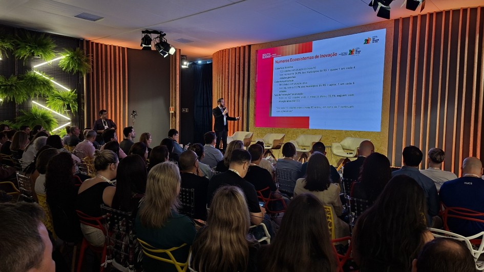 Foto mostra palestrante no palco mostrando dados numa tela, enquanto plateia acompanha apresenta&ccedil;&atilde;o.