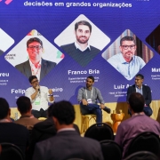Painel de debate em evento corporativo, com cinco participantes sentados no palco e plateia ao fundo. Um tel&atilde;o azul exibe o tema sobre decis&otilde;es em grandes organiza&ccedil;&otilde;es, com nomes e informa&ccedil;&otilde;es dos painelistas.