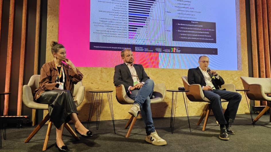 Painel de discuss&atilde;o em evento institucional, com tr&ecirc;s participantes sentados em poltronas no palco. Ao fundo, um tel&atilde;o exibe gr&aacute;ficos e informa&ccedil;&otilde;es sobre startups. 