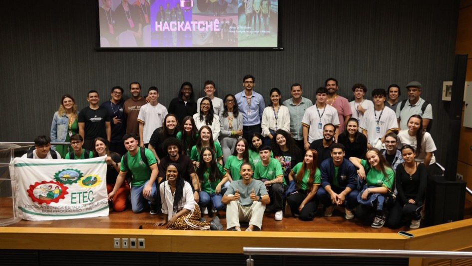 Esta &eacute; uma descri&ccedil;&atilde;o detalhada da imagem para fins de acessibilidade:

A fotografia mostra um grande grupo de jovens e alguns adultos reunidos no palco de um audit&oacute;rio para o an&uacute;ncio das equipes finalistas do HackaTch&ecirc; 2026.

Elementos Centrais e Composi&ccedil;&atilde;o
O Grupo: Cerca de 30 pessoas posam para a foto. O grupo &eacute; diversificado e demonstra entusiasmo. Muitos jovens vestem camisetas verdes com o logotipo do evento, enquanto outros usam roupas casuais. Eles est&atilde;o organizados em diferentes n&iacute;veis: alguns em p&eacute; ao fundo, outros sentados ou agachados &agrave; frente.

Destaque Visual (Esquerda): Dois jovens seguram uma bandeira branca da ETEC (Escola T&eacute;cnica Estadual Celeiro), de Bom Progresso - RS. A bandeira cont&eacute;m ilustra&ccedil;&otilde;es coloridas que remetem &agrave; agricultura e ao ensino t&eacute;cnico.

Proje&ccedil;&atilde;o ao Fundo: No alto, uma tela de proje&ccedil;&atilde;o exibe a identidade visual do evento com o nome "HACKATCH&Ecirc;" em letras garrafais brancas, sob a imagem de uma l&acirc;mpada estilizada (s&iacute;mbolo de ideia/inova&ccedil;&atilde;o) feita de circuitos de luz roxa.

Ambiente do Audit&oacute;rio
Palco: O piso &eacute; de madeira clara (tablado), e a parte frontal do palco tem um acabamento em madeira com detalhes met&aacute;licos.

Paredes: O fundo do palco &eacute; composto por pain&eacute;is ac&uacute;sticos ripados em tom cinza-escuro, conferindo um aspecto moderno e profissional ao ambiente.

Ilumina&ccedil;&atilde;o: A luz &eacute; focada no grupo, destacando as express&otilde;es de alegria e conquista dos participantes.

Atmosfera
A imagem transmite um clima de celebra&ccedil;&atilde;o, inova&ccedil;&atilde;o e protagonismo juvenil. &Eacute; um registro de encerramento de uma etapa importante, onde o esp&iacute;rito de equipe e o orgulho pela representa&ccedil;&atilde;o de suas escolas s&atilde;o evidentes.