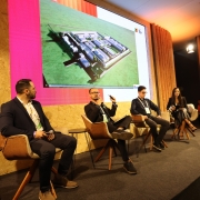 Foto mostra palestrante sentado, olhando para tela onde h&aacute; uma foto de um projeto de uma unidade prisional. Outros tr&ecirc;s palestrantes acompanham a fala, tamb&eacute;m sentados. 