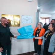 A fotografia registra o momento solene e festivo do descerramento de duas placas de inaugura&ccedil;&atilde;o em uma unidade de sa&uacute;de, com a presen&ccedil;a de autoridades estaduais e municipais.

A A&ccedil;&atilde;o Central (Descerramento)
Pessoas: Quatro pessoas est&atilde;o em destaque &agrave; frente de uma parede branca.

Esquerda: Um homem de barba, vestindo uma camisa preta e cal&ccedil;a escura, sorri e segura uma das pontas de um tecido azul-turquesa que cobria as placas.

Direita: Duas mulheres e um homem est&atilde;o em p&eacute; ao lado dele, tamb&eacute;m sorrindo e participando do descerramento.

A mulher ao centro veste um blazer laranja vibrante sobre uma blusa preta, com um crach&aacute; colorido no pesco&ccedil;o.

A mulher mais &agrave; direita veste um blazer cinza-claro e segura a outra ponta do tecido azul.

O Tecido: Um pano azul-turquesa claro, que cobria as placas, est&aacute; sendo puxado e dobra-se no ar, revelando as placas.

As Placas (Fundo)
Placa da Esquerda: Uma placa dourada retangular e reluzente. O texto &eacute; mais denso e formal, incluindo logotipos do HOSPITAL BOM PASTOR DE GRAVATA&Iacute; e men&ccedil;&otilde;es a atos municipais e estaduais de coopera&ccedil;&atilde;o.

Placa da Direita: Uma placa branca emoldurada de forma mais simples. &Eacute; a placa principal de inaugura&ccedil;&atilde;o, com o bras&atilde;o do Estado do Rio Grande do Sul no topo. O texto principal &eacute;: "INAUGURA&Ccedil;&Atilde;O DO NOVO TELHADO DO HOSPITAL", especificando que o investimento &eacute; do Programa Avan&ccedil;ar na Sa&uacute;de do Governo do Estado. Abaixo, l&ecirc;-se o nome do governador "Eduardo Leite" e de outros secret&aacute;rios estaduais.

Ambiente e Outras Pessoas
Ambiente: A parede &eacute; branca com um barrado de m&aacute;rmore esverdeado na base. Ao fundo, v&ecirc;-se um corredor amplo com piso de cimento, colunas brancas e janelas com cortinas, al&eacute;m de mais pessoas que observam a cerim&ocirc;nia. A ilumina&ccedil;&atilde;o &eacute; de teto, conferindo um tom claro e natural &agrave; cena.

P&uacute;blico: &Agrave; direita, atr&aacute;s da mulher de cinza, um homem de terno preto e outra pessoa assistem &agrave; cena. Mais ao fundo, distinguem-se outras silhuetas e um extintor de inc&ecirc;ndio na parede.

Atmosfera
A cena transmite alegria, orgulho e coopera&ccedil;&atilde;o entre as diferentes esferas de governo para a melhoria de uma infraestrutura p&uacute;blica de sa&uacute;de. &Eacute; um registro de uma conquista entregue &agrave; comunidade.