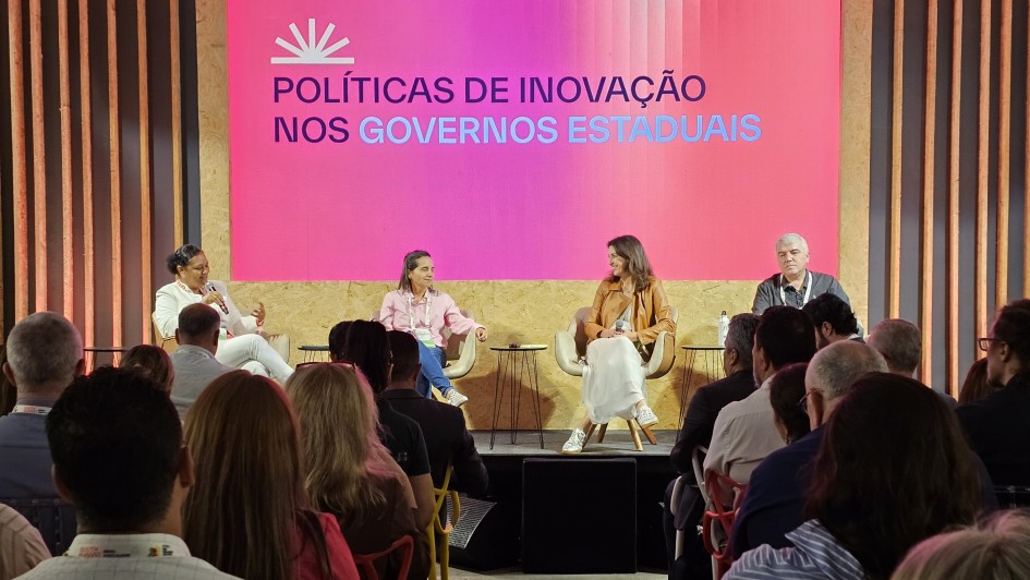 Painel de debate em evento institucional, com quatro participantes sentados em poltronas no palco e plateia ocupando fileiras de cadeiras &agrave; frente. Ao fundo, um grande tel&atilde;o rosa exibe o t&iacute;tulo &ldquo;Pol&iacute;ticas de Inova&ccedil;&atilde;o nos Governos Estaduais&rdquo;, com cenografia em pain&eacute;is de madeira e ilumina&ccedil;&atilde;o c&ecirc;nica.