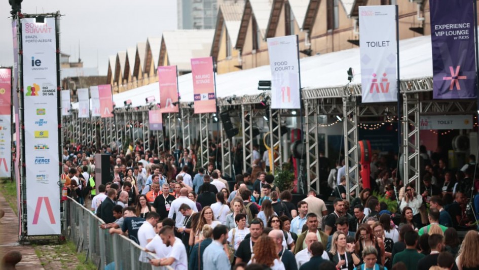 A fotografia a&eacute;rea, capturada em um &acirc;ngulo levemente elevado, mostra a intensa movimenta&ccedil;&atilde;o de p&uacute;blico no Cais Embarcadero, em Porto Alegre, durante o South Summit Brazil 2026.

O P&uacute;blico e a Movimenta&ccedil;&atilde;o
Multid&atilde;o: Centenas de pessoas de perfis diversos circulam por um amplo corredor a c&eacute;u aberto. A maioria veste roupas casuais ou profissionais (estilo business casual) e muitos portam crach&aacute;s oficiais do evento pendurados no pesco&ccedil;o. O clima &eacute; de grande efervesc&ecirc;ncia e circula&ccedil;&atilde;o constante.

Corredor Principal: O fluxo de pessoas ocorre entre uma grade de prote&ccedil;&atilde;o &agrave; esquerda (que separa a &aacute;rea do Gua&iacute;ba) e uma estrutura de tendas brancas e estandes &agrave; direita.

Estruturas e Sinaliza&ccedil;&atilde;o
Banners e Totens: Diversas bandeiras verticais e totens de sinaliza&ccedil;&atilde;o em tons de branco, rosa e azul est&atilde;o espalhados pelo trajeto, orientando o p&uacute;blico para &aacute;reas como:

Food Court 2 (Pra&ccedil;a de alimenta&ccedil;&atilde;o)

Music Stage (Palco de m&uacute;sica)

Toilet (Banheiros)

Experience VIP Lounge by PUCRS

Patroc&iacute;nios e Parcerias: Um grande totem &agrave; esquerda exibe os logotipos dos principais realizadores e parceiros, com destaque para o Governo do Estado do Rio Grande do Sul, SEBRAE, FIERGS e a prefeitura de Porto Alegre.

Arquitetura do Cais: Ao fundo, alinham-se os hist&oacute;ricos telhados em formato de "serra" (shed) dos armaz&eacute;ns do Cais, que contrastam com as estruturas met&aacute;licas modernas e as tendas tempor&aacute;rias montadas para o evento tecnol&oacute;gico.

Ambiente e Composi&ccedil;&atilde;o
C&eacute;u e Ilumina&ccedil;&atilde;o: O dia parece nublado, com uma luz difusa que ilumina uniformemente a multid&atilde;o sem criar sombras fortes.

Perspectiva: A profundidade de campo mostra que o evento se estende por uma longa dist&acirc;ncia ao longo da orla, enfatizando a magnitude da infraestrutura montada.

Atmosfera
A imagem transmite uma sensa&ccedil;&atilde;o de vitalidade, conex&atilde;o e escala, posicionando o Rio Grande do Sul como um polo vibrante de inova&ccedil;&atilde;o, tecnologia e grandes encontros globais.