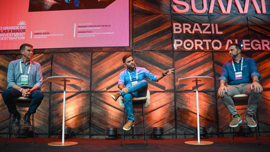 Palco do South Summit Brazil &ndash; Porto Alegre, com tr&ecirc;s participantes distribu&iacute;dos em assentos altos e mesas individuais. Ao fundo, a identidade visual do evento aparece em letras grandes, integrada &agrave; cenografia de madeira e luzes direcionadas.