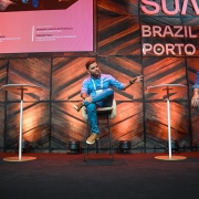Palco do South Summit Brazil &ndash; Porto Alegre, com tr&ecirc;s participantes distribu&iacute;dos em assentos altos e mesas individuais. Ao fundo, a identidade visual do evento aparece em letras grandes, integrada &agrave; cenografia de madeira e luzes direcionadas.