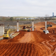 Imagem de uma grande &aacute;rea em obras, com maquin&aacute;rio como retroescavadeiras e ca&ccedil;ambas circulando na &aacute;rea. No centro, tr&ecirc;s grandes pilares de concreto