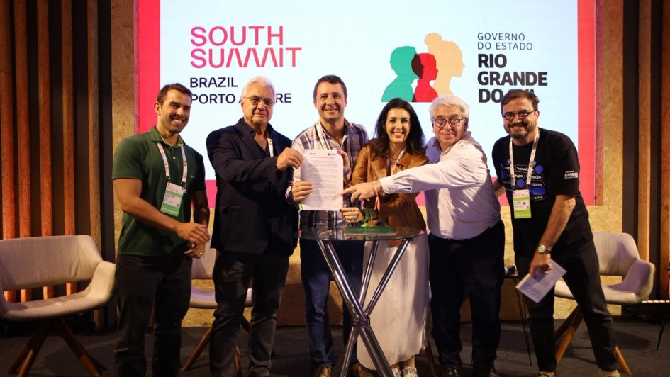Um grupo de seis pessoas se posiciona ao redor de uma pequena mesa segurando um documento. Ao fundo, um painel com as logo marcas do South Summit Brazil e do governo do RS.