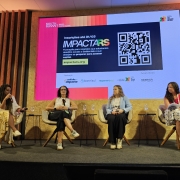 Quatro mulheres sentadas em cadeiras dispostas em um palco. Aoo fundo, um banner com a logomarca da ImpactaRS
