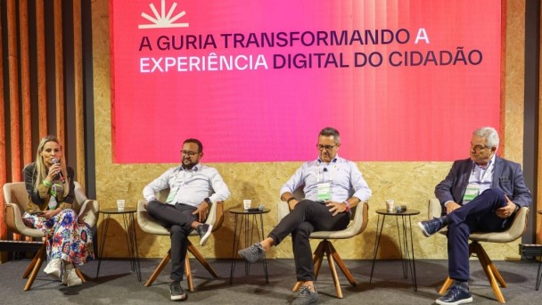 A foto registra um painel no South Summit Brazil, com quatro participantes sentados em poltronas diante de um grande tel&atilde;o rosa que exibe o texto: &ldquo;A guria transformando a experi&ecirc;ncia digital do cidad&atilde;o&rdquo;. O palco tem decora&ccedil;&atilde;o em madeira vertical e ilumina&ccedil;&atilde;o direcionada, enquanto a plateia assiste ao debate nas primeiras fileiras.