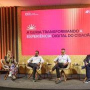 A foto registra um painel no South Summit Brazil, com quatro participantes sentados em poltronas diante de um grande tel&atilde;o rosa que exibe o texto: &ldquo;A guria transformando a experi&ecirc;ncia digital do cidad&atilde;o&rdquo;. O palco tem decora&ccedil;&atilde;o em madeira vertical e ilumina&ccedil;&atilde;o direcionada, enquanto a plateia assiste ao debate nas primeiras fileiras.