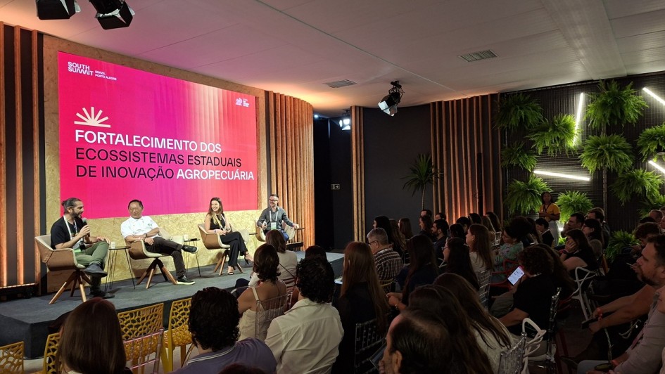 Esta &eacute; uma descri&ccedil;&atilde;o detalhada da imagem para fins de acessibilidade:

A fotografia registra um painel de discuss&atilde;o durante o South Summit Brazil 2026, focado no setor prim&aacute;rio e inova&ccedil;&atilde;o.

O Palco e a Apresenta&ccedil;&atilde;o
Painelistas: Quatro pessoas (tr&ecirc;s homens e uma mulher) est&atilde;o sentadas em poltronas girat&oacute;rias de cor clara sobre um palco baixo. Eles est&atilde;o dispostos em semic&iacute;rculo, voltados uns para os outros e para a plateia, em um clima de conversa informal.

O Tel&atilde;o: Um grande painel de LED ao fundo exibe um slide com fundo rosa vibrante e letras brancas, onde se l&ecirc;: "FORTALECIMENTO DOS ECOSSISTEMAS ESTADUAIS DE INOVA&Ccedil;&Atilde;O AGROPECU&Aacute;RIA". No canto superior esquerdo do tel&atilde;o, aparece o logotipo do South Summit Brazil Porto Alegre.

Cenografia: O palco &eacute; emoldurado por pain&eacute;is de madeira clara e ripas verticais que criam um efeito de luz e sombra.

O Ambiente e o P&uacute;blico
Audit&oacute;rio: A plateia est&aacute; lotada, com dezenas de pessoas sentadas em cadeiras transparentes e coloridas (amarelas e vermelhas). O p&uacute;blico est&aacute; de costas para a c&acirc;mera, com o olhar atento ao palco.

Decora&ccedil;&atilde;o Lateral: &Agrave; direita da imagem, destaca-se uma parede decorada com samambaias verdes dispostas em uma grade met&aacute;lica, iluminada por tubos de LED branco em formatos geom&eacute;tricos, similar &agrave; cenografia de outros pain&eacute;is do evento.

Ilumina&ccedil;&atilde;o: O ambiente possui uma ilumina&ccedil;&atilde;o c&ecirc;nica sofisticada, com refletores de luz negra e branca pendurados no teto, criando um contraste moderno entre o palco iluminado e a plateia na penumbra.

Atmosfera
A cena transmite uma sensa&ccedil;&atilde;o de conhecimento compartilhado e foco tecnol&oacute;gico. A imagem refor&ccedil;a a import&acirc;ncia do agroneg&oacute;cio dentro do ecossistema de inova&ccedil;&atilde;o global, unindo especialistas para debater o futuro do setor no Rio Grande do Sul.