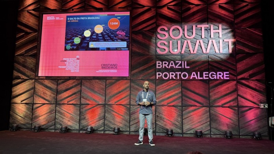 Homem em um grande palco de p&eacute;, em palestra. Atr&aacute;s dele, uma parede decorada, com luzes e a logomarca South Summit Brazil