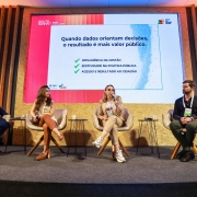 A imagem mostra um painel no South Summit Brazil, com quatro participantes sentados em poltronas diante de um tel&atilde;o grande que exibe o texto: &ldquo;Quando dados orientam decis&otilde;es, o resultado &eacute; mais valor p&uacute;blico&rdquo; acompanhado de tr&ecirc;s t&oacute;picos listados com marcas de sele&ccedil;&atilde;o. 
