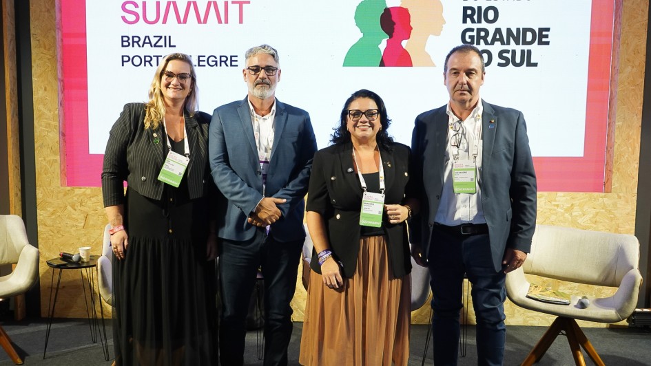 A imagem mostra quatro pessoas alinhadas no palco de um evento, posicionadas diante de um tel&atilde;o com a identidade visual do South Summit Brazil &ndash; Porto Alegre e de um programa institucional do governo estadual. Todas est&atilde;o em p&eacute;, com crach&aacute;s de identifica&ccedil;&atilde;o, e atr&aacute;s delas h&aacute; poltronas claras utilizadas no painel. 