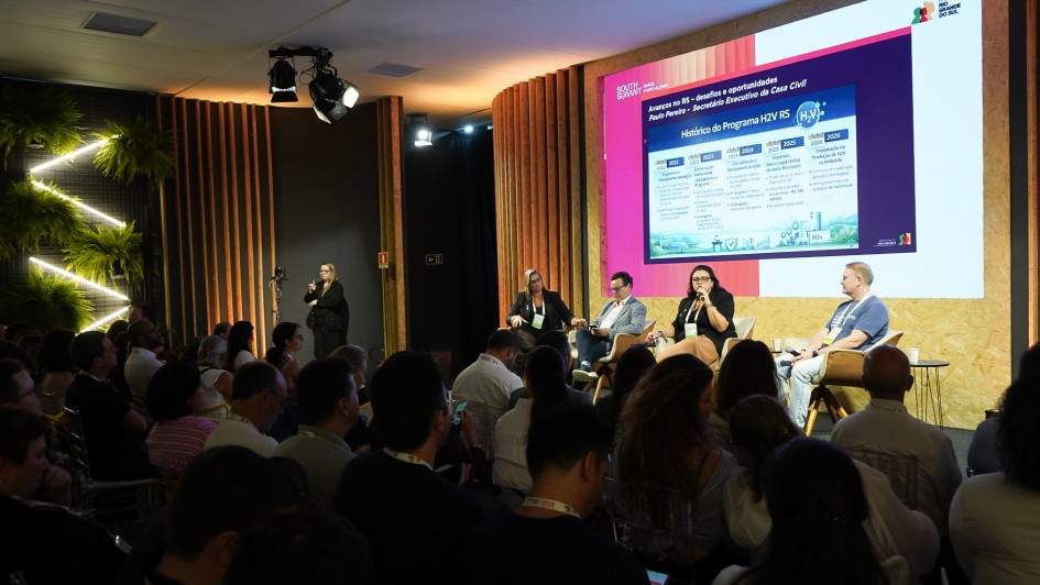 A imagem mostra um painel em andamento, com quatro pessoas sentadas em poltronas no palco. &Agrave; frente delas, a plateia ocupa cadeiras organizadas em fileiras. No fundo, um tel&atilde;o exibe informa&ccedil;&otilde;es do Programa H2V RS, incluindo uma linha do tempo e &iacute;cones ilustrativos. 