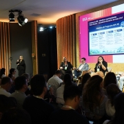 A imagem mostra um painel em andamento, com quatro pessoas sentadas em poltronas no palco. &Agrave; frente delas, a plateia ocupa cadeiras organizadas em fileiras. No fundo, um tel&atilde;o exibe informa&ccedil;&otilde;es do Programa H2V RS, incluindo uma linha do tempo e &iacute;cones ilustrativos. 