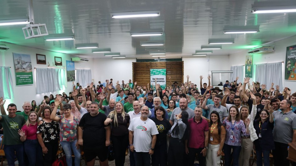 A imagem mostra um grande grupo reunido em um sal&atilde;o amplo, iluminado por luz artificial branca vinda do teto. As pessoas est&atilde;o distribu&iacute;das em p&eacute; por todo o espa&ccedil;o, muitas delas com os bra&ccedil;os erguidos.