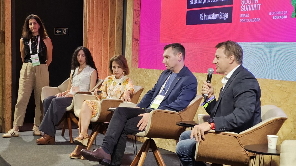 A imagem mostra um painel realizado em um palco de evento, com quatro pessoas sentadas em poltronas posicionadas em semic&iacute;rculo. &Agrave; esquerda, outra pessoa permanece em p&eacute;, acompanhando a conversa. Ao fundo, um tel&atilde;o exibe informa&ccedil;&otilde;es do South Summit Brazil e detalhes da programa&ccedil;&atilde;o.