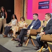 A imagem mostra um painel realizado em um palco de evento, com quatro pessoas sentadas em poltronas posicionadas em semic&iacute;rculo. &Agrave; esquerda, outra pessoa permanece em p&eacute;, acompanhando a conversa. Ao fundo, um tel&atilde;o exibe informa&ccedil;&otilde;es do South Summit Brazil e detalhes da programa&ccedil;&atilde;o.