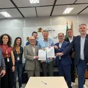 Grupo de nove pessoas lado a lado seguram duas folhas em uma sala. &Agrave; frente, uma mesa vazia com uma caneta. 