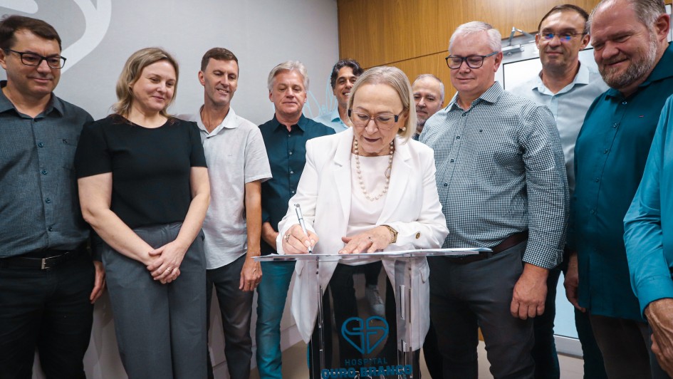 A imagem mostra um grupo de cerca de dez pessoas reunidas em um ambiente interno moderno para um ato oficial de assinatura. O foco central est&aacute; em uma mulher de meia-idade, vestindo um blazer branco, que est&aacute; inclinada sobre um p&uacute;lpito de acr&iacute;lico transparente assinando um documento.

Detalhes da Cena
A Figura Central: Uma mulher de pele clara e cabelos loiros curtos. Ela usa &oacute;culos de arma&ccedil;&atilde;o fina, um colar de p&eacute;rolas e um blazer branco sobre uma blusa clara. Ela segura uma caneta e est&aacute; concentrada no documento sobre o p&uacute;lpito.

O Grupo: Ao redor dela, homens e mulheres de diversas idades observam o ato. Eles est&atilde;o vestidos de forma casual-social (camisas de bot&atilde;o, camisetas polo e cal&ccedil;as sociais). As express&otilde;es variam entre sorrisos discretos e olhares atentos.

O P&uacute;lpito: &Eacute; feito de material transparente (acr&iacute;lico). Na parte frontal, h&aacute; um logotipo azul composto por dois cora&ccedil;&otilde;es entrela&ccedil;ados que formam uma figura estilizada, seguido pelo texto "HOSPITAL OURO BRANCO - EXCEL&Ecirc;NCIA EM SA&Uacute;DE".

O Ambiente: O fundo consiste em uma parede cinza clara &agrave; esquerda, com uma parte de um grande logotipo circular branco, e uma parede com pain&eacute;is de madeira clara &agrave; direita. A ilumina&ccedil;&atilde;o &eacute; direta e clara, t&iacute;pica de um escrit&oacute;rio ou ambiente hospitalar moderno.

Composi&ccedil;&atilde;o
A foto &eacute; tirada de um &acirc;ngulo ligeiramente baixo, o que d&aacute; import&acirc;ncia ao momento da assinatura. As pessoas est&atilde;o dispostas em semic&iacute;rculo atr&aacute;s e aos lados da autoridade central, criando uma sensa&ccedil;&atilde;o de uni&atilde;o e celebra&ccedil;&atilde;o de uma conquista institucional.