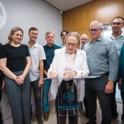 A imagem mostra um grupo de cerca de dez pessoas reunidas em um ambiente interno moderno para um ato oficial de assinatura. O foco central est&aacute; em uma mulher de meia-idade, vestindo um blazer branco, que est&aacute; inclinada sobre um p&uacute;lpito de acr&iacute;lico transparente assinando um documento.

Detalhes da Cena
A Figura Central: Uma mulher de pele clara e cabelos loiros curtos. Ela usa &oacute;culos de arma&ccedil;&atilde;o fina, um colar de p&eacute;rolas e um blazer branco sobre uma blusa clara. Ela segura uma caneta e est&aacute; concentrada no documento sobre o p&uacute;lpito.

O Grupo: Ao redor dela, homens e mulheres de diversas idades observam o ato. Eles est&atilde;o vestidos de forma casual-social (camisas de bot&atilde;o, camisetas polo e cal&ccedil;as sociais). As express&otilde;es variam entre sorrisos discretos e olhares atentos.

O P&uacute;lpito: &Eacute; feito de material transparente (acr&iacute;lico). Na parte frontal, h&aacute; um logotipo azul composto por dois cora&ccedil;&otilde;es entrela&ccedil;ados que formam uma figura estilizada, seguido pelo texto "HOSPITAL OURO BRANCO - EXCEL&Ecirc;NCIA EM SA&Uacute;DE".

O Ambiente: O fundo consiste em uma parede cinza clara &agrave; esquerda, com uma parte de um grande logotipo circular branco, e uma parede com pain&eacute;is de madeira clara &agrave; direita. A ilumina&ccedil;&atilde;o &eacute; direta e clara, t&iacute;pica de um escrit&oacute;rio ou ambiente hospitalar moderno.

Composi&ccedil;&atilde;o
A foto &eacute; tirada de um &acirc;ngulo ligeiramente baixo, o que d&aacute; import&acirc;ncia ao momento da assinatura. As pessoas est&atilde;o dispostas em semic&iacute;rculo atr&aacute;s e aos lados da autoridade central, criando uma sensa&ccedil;&atilde;o de uni&atilde;o e celebra&ccedil;&atilde;o de uma conquista institucional.