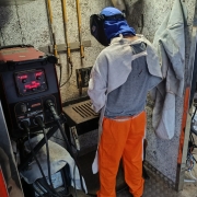 A imagem mostra uma pessoa de costas realizando um trabalho t&eacute;cnico de soldagem em um ambiente industrial ou de oficina. A pessoa est&aacute; equipada com vestimentas de prote&ccedil;&atilde;o e opera um equipamento profissional.

Detalhes do Indiv&iacute;duo e Equipamento
Vestimenta: A pessoa veste uma cal&ccedil;a laranja vibrante e uma camiseta cinza. Por cima, utiliza Equipamentos de Prote&ccedil;&atilde;o Individual (EPIs), incluindo um avental de raspa cinza (couro protetor) que cobre os ombros e as costas, e uma balaclava azul que cobre toda a cabe&ccedil;a e pesco&ccedil;o sob a m&aacute;scara de solda preta, que est&aacute; levantada no momento.

M&aacute;quina de Solda: &Agrave; esquerda, h&aacute; uma m&aacute;quina de solda industrial robusta de cor preta e vermelha, modelo Lincoln Electric C300 CE. O painel digital exibe n&uacute;meros em vermelho (indicando 114 e 20.0) e diversas luzes de controle acesas. Cabos pretos grossos conectam a m&aacute;quina &agrave; &aacute;rea de trabalho.

&Aacute;rea de Trabalho: &Agrave; frente da pessoa, h&aacute; uma bancada de metal com uma grelha preta onde o trabalho de soldagem &eacute; realizado. A parede ao fundo tem uma textura rugosa e clara, com tubula&ccedil;&otilde;es e suportes met&aacute;licos fixados.

Ambiente e Contexto
Cen&aacute;rio: O local parece ser uma unidade m&oacute;vel ou uma oficina especializada, com paredes revestidas de material isolante. &Agrave; direita, pendurados na parede, veem-se outros aventais de prote&ccedil;&atilde;o de couro cinza. O ch&atilde;o &eacute; de metal antiderrapante (estilo "piso de &ocirc;nibus" ou chapa xadrez).

Ilumina&ccedil;&atilde;o: A luz &eacute; funcional e focada na &aacute;rea de opera&ccedil;&atilde;o, destacando o contraste entre o laranja da cal&ccedil;a e os tons industriais de cinza e preto do restante da cena.