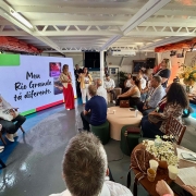 A imagem mostra um encontro realizado em um espa&ccedil;o interno iluminado por luz natural lateral. Um tel&atilde;o grande exibe a frase &ldquo;Meu Rio Grande t&aacute; diferente.&rdquo;, acompanhada da identidade visual do South Summit Brazil. Pessoas est&atilde;o distribu&iacute;das em assentos baixos e mesas de apoio, enquanto duas pessoas ficam &agrave; frente do tel&atilde;o conduzindo a apresenta&ccedil;&atilde;o.