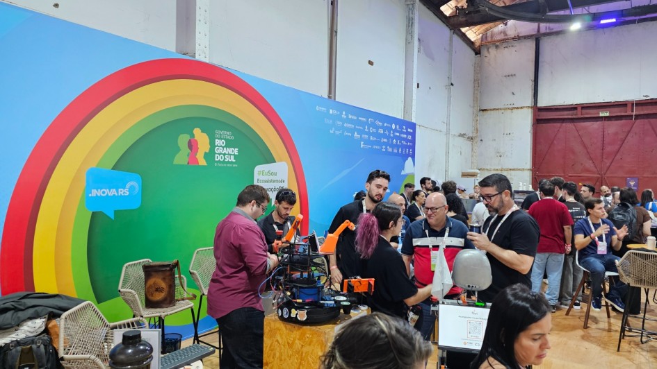 A imagem mostra um estande de inova&ccedil;&atilde;o tecnol&oacute;gica em um pavilh&atilde;o amplo, com caracter&iacute;sticas industriais. Diversas pessoas circulam pelo espa&ccedil;o, interagindo com prot&oacute;tipos e equipamentos eletr&ocirc;nicos dispostos sobre balc&otilde;es de madeira.

Detalhes do Estande
Painel de Fundo: Um grande painel azul ocupa o lado esquerdo. Ele apresenta um arco colorido (em tons de amarelo, laranja e vermelho) que circunda o logotipo do Governo do Estado do Rio Grande do Sul. &Agrave; esquerda, h&aacute; um bal&atilde;o de fala azul com a inscri&ccedil;&atilde;o ".INOVA RS".

Intera&ccedil;&atilde;o e Tecnologia: No centro, um grupo de jovens (homens e uma mulher com cabelo rosa) manipula um prot&oacute;tipo rob&oacute;tico complexo com pe&ccedil;as laranjas, montado sobre uma base de madeira compensada (OSB). H&aacute; fios, placas de circuito e pequenos monitores vis&iacute;veis.

Equipamentos: &Agrave; direita, em primeiro plano, v&ecirc;-se um terminal de computador com teclado e um pequeno busto prateado de rob&ocirc; ao lado. Um QR Code est&aacute; vis&iacute;vel na tela do monitor.

Ambiente e P&uacute;blico
Cen&aacute;rio: O evento ocorre em um galp&atilde;o com p&eacute;-direito alto, paredes brancas desgastadas e vigas met&aacute;licas expostas. Ao fundo, uma grande porta met&aacute;lica vermelha e trilhos no teto refor&ccedil;am a est&eacute;tica industrial do local.

Pessoas: O p&uacute;blico &eacute; diversificado, vestindo roupas casuais. Algumas pessoas usam crach&aacute;s no pesco&ccedil;o. No lado direito, h&aacute; pessoas sentadas em banquetas altas de palha conversando, criando um ambiente de networking e troca de conhecimento.

Composi&ccedil;&atilde;o
A foto &eacute; ampla e bem iluminada, capturando o dinamismo do evento. A profundidade de campo permite ver desde os detalhes t&eacute;cnicos do rob&ocirc; em primeiro plano at&eacute; a movimenta&ccedil;&atilde;o da multid&atilde;o ao fundo.