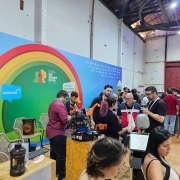 A imagem mostra um estande de inova&ccedil;&atilde;o tecnol&oacute;gica em um pavilh&atilde;o amplo, com caracter&iacute;sticas industriais. Diversas pessoas circulam pelo espa&ccedil;o, interagindo com prot&oacute;tipos e equipamentos eletr&ocirc;nicos dispostos sobre balc&otilde;es de madeira.

Detalhes do Estande
Painel de Fundo: Um grande painel azul ocupa o lado esquerdo. Ele apresenta um arco colorido (em tons de amarelo, laranja e vermelho) que circunda o logotipo do Governo do Estado do Rio Grande do Sul. &Agrave; esquerda, h&aacute; um bal&atilde;o de fala azul com a inscri&ccedil;&atilde;o ".INOVA RS".

Intera&ccedil;&atilde;o e Tecnologia: No centro, um grupo de jovens (homens e uma mulher com cabelo rosa) manipula um prot&oacute;tipo rob&oacute;tico complexo com pe&ccedil;as laranjas, montado sobre uma base de madeira compensada (OSB). H&aacute; fios, placas de circuito e pequenos monitores vis&iacute;veis.

Equipamentos: &Agrave; direita, em primeiro plano, v&ecirc;-se um terminal de computador com teclado e um pequeno busto prateado de rob&ocirc; ao lado. Um QR Code est&aacute; vis&iacute;vel na tela do monitor.

Ambiente e P&uacute;blico
Cen&aacute;rio: O evento ocorre em um galp&atilde;o com p&eacute;-direito alto, paredes brancas desgastadas e vigas met&aacute;licas expostas. Ao fundo, uma grande porta met&aacute;lica vermelha e trilhos no teto refor&ccedil;am a est&eacute;tica industrial do local.

Pessoas: O p&uacute;blico &eacute; diversificado, vestindo roupas casuais. Algumas pessoas usam crach&aacute;s no pesco&ccedil;o. No lado direito, h&aacute; pessoas sentadas em banquetas altas de palha conversando, criando um ambiente de networking e troca de conhecimento.

Composi&ccedil;&atilde;o
A foto &eacute; ampla e bem iluminada, capturando o dinamismo do evento. A profundidade de campo permite ver desde os detalhes t&eacute;cnicos do rob&ocirc; em primeiro plano at&eacute; a movimenta&ccedil;&atilde;o da multid&atilde;o ao fundo.