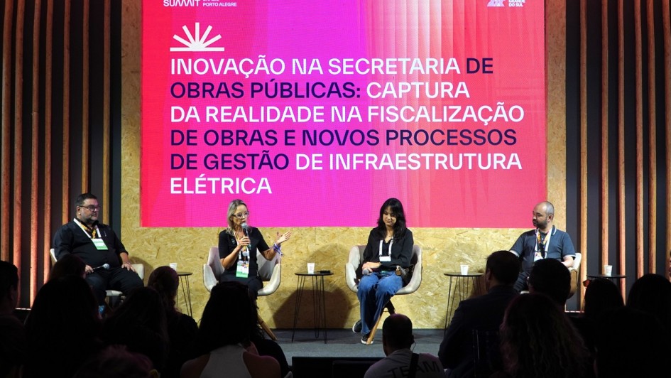 A imagem registra um painel de debate realizado em um palco do evento South Summit Brazil. Quatro palestrantes est&atilde;o sentados em poltronas bege, dispostos em linha, diante de um grande tel&atilde;o iluminado. A silhueta de parte da plateia &eacute; vis&iacute;vel em primeiro plano, na parte inferior da foto.

Detalhes do Palco e Palestrantes
Os Participantes: Da esquerda para a direita, h&aacute; um homem de barba e camisa preta; uma mulher loira de &oacute;culos e blusa preta que fala ao microfone e gesticula com a m&atilde;o esquerda; uma mulher de cabelos escuros e cal&ccedil;a jeans; e um homem de barba e camiseta polo cinza. Todos utilizam crach&aacute;s de identifica&ccedil;&atilde;o do evento.

O Tel&atilde;o: Possui um fundo em degrad&ecirc; vibrante que vai do rosa ao magenta. No topo, l&ecirc;-se "SUMMIT PORTO ALEGRE". O texto central, em letras brancas mai&uacute;sculas, diz: "INOVA&Ccedil;&Atilde;O NA SECRETARIA DE OBRAS P&Uacute;BLICAS: CAPTURA DA REALIDADE NA FISCALIZA&Ccedil;&Atilde;O DE OBRAS E NOVOS PROCESSOS DE GEST&Atilde;O DE INFRAESTRUTURA EL&Eacute;TRICA".

Mobili&aacute;rio: Entre os palestrantes, h&aacute; pequenas mesas de apoio pretas com copos de &aacute;gua descart&aacute;veis.

Cen&aacute;rio e Composi&ccedil;&atilde;o
Decora&ccedil;&atilde;o: O palco &eacute; revestido com placas de madeira clara (OSB) na parte inferior. Nas laterais, h&aacute; uma estrutura decorativa composta por ripas de madeira verticais que se estendem at&eacute; o teto, criando um efeito de profundidade.

Ilumina&ccedil;&atilde;o: A luz principal prov&eacute;m do tel&atilde;o e de refletores direcionados aos palestrantes, destacando-os contra o fundo colorido. A plateia est&aacute; mergulhada em sombra, com apenas as silhuetas de cabe&ccedil;as e ombros vis&iacute;veis.