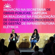 A imagem registra um painel de debate realizado em um palco do evento South Summit Brazil. Quatro palestrantes est&atilde;o sentados em poltronas bege, dispostos em linha, diante de um grande tel&atilde;o iluminado. A silhueta de parte da plateia &eacute; vis&iacute;vel em primeiro plano, na parte inferior da foto.

Detalhes do Palco e Palestrantes
Os Participantes: Da esquerda para a direita, h&aacute; um homem de barba e camisa preta; uma mulher loira de &oacute;culos e blusa preta que fala ao microfone e gesticula com a m&atilde;o esquerda; uma mulher de cabelos escuros e cal&ccedil;a jeans; e um homem de barba e camiseta polo cinza. Todos utilizam crach&aacute;s de identifica&ccedil;&atilde;o do evento.

O Tel&atilde;o: Possui um fundo em degrad&ecirc; vibrante que vai do rosa ao magenta. No topo, l&ecirc;-se "SUMMIT PORTO ALEGRE". O texto central, em letras brancas mai&uacute;sculas, diz: "INOVA&Ccedil;&Atilde;O NA SECRETARIA DE OBRAS P&Uacute;BLICAS: CAPTURA DA REALIDADE NA FISCALIZA&Ccedil;&Atilde;O DE OBRAS E NOVOS PROCESSOS DE GEST&Atilde;O DE INFRAESTRUTURA EL&Eacute;TRICA".

Mobili&aacute;rio: Entre os palestrantes, h&aacute; pequenas mesas de apoio pretas com copos de &aacute;gua descart&aacute;veis.

Cen&aacute;rio e Composi&ccedil;&atilde;o
Decora&ccedil;&atilde;o: O palco &eacute; revestido com placas de madeira clara (OSB) na parte inferior. Nas laterais, h&aacute; uma estrutura decorativa composta por ripas de madeira verticais que se estendem at&eacute; o teto, criando um efeito de profundidade.

Ilumina&ccedil;&atilde;o: A luz principal prov&eacute;m do tel&atilde;o e de refletores direcionados aos palestrantes, destacando-os contra o fundo colorido. A plateia est&aacute; mergulhada em sombra, com apenas as silhuetas de cabe&ccedil;as e ombros vis&iacute;veis.