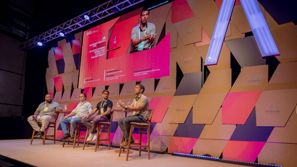 Esta &eacute; uma descri&ccedil;&atilde;o detalhada da imagem para fins de acessibilidade:

Vis&atilde;o Geral
A imagem mostra um painel de debate no evento South Summit Brazil. Quatro homens est&atilde;o sentados em banquetas altas de madeira sobre um palco, participando de uma discuss&atilde;o. Ao fundo, um cen&aacute;rio din&acirc;mico e um grande tel&atilde;o comp&otilde;em o ambiente.

Detalhes do Palco e Palestrantes
Os Participantes: Da esquerda para a direita, o primeiro homem veste uma camisa polo clara e cal&ccedil;a bege; o segundo, uma camisa social azul clara e jeans; o terceiro, uma camiseta preta; e o quarto, que est&aacute; falando e gesticulando no momento, veste uma camisa polo bege e cal&ccedil;a escura. Todos utilizam crach&aacute;s do evento.

O Tel&atilde;o: Exibe uma transmiss&atilde;o ao vivo em close-up do palestrante que est&aacute; falando. No lado esquerdo do tel&atilde;o, h&aacute; um texto em ingl&ecirc;s sobre fundo rosa: "BLENDED FINANCE FOR RESILIENT DEVELOPMENT: THE ROLE OF DEVELOPMENT BANKS AND CATALYTIC PHILANTHROPY". Abaixo, os nomes dos participantes e suas institui&ccedil;&otilde;es s&atilde;o listados em letras menores.

Cen&aacute;rio de Fundo: A parede atr&aacute;s dos palestrantes &eacute; decorada com pain&eacute;is geom&eacute;tricos sobrepostos em tons de madeira natural, rosa, azul escuro e cinza. Algumas pe&ccedil;as possuem o logotipo do evento e do Governo do Estado do Rio Grande do Sul. Uma grande estrutura luminosa em formato de "V" invertido ou parte de um "A" se destaca no lado direito.

Composi&ccedil;&atilde;o e Ilumina&ccedil;&atilde;o
Ambiente: O palco tem um acabamento em carpete bege claro. Uma fileira de luzes LED no ch&atilde;o, na borda frontal do palco, emite uma luz azulada. No topo da estrutura, refletores de palco iluminam a cena.

Perspectiva: A foto &eacute; tirada de um &acirc;ngulo lateral e ligeiramente de baixo, capturando a extens&atilde;o do palco e a modernidade da cenografia.