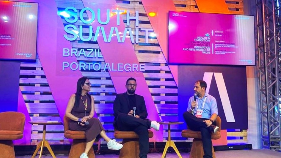 Esta &eacute; uma descri&ccedil;&atilde;o detalhada da imagem para fins de acessibilidade:

Vis&atilde;o Geral
A imagem apresenta um painel de debate no evento South Summit Brazil 2026. Tr&ecirc;s palestrantes (uma mulher e dois homens) est&atilde;o sentados em poltronas modernas sobre um palco, engajados em uma conversa. O cen&aacute;rio ao fundo &eacute; vibrante e utiliza elementos de madeira e ilumina&ccedil;&atilde;o colorida.

Detalhes do Palco e Palestrantes
Os Participantes: &Agrave; esquerda, uma mulher de cabelos escuros e &oacute;culos, vestindo um vestido marrom e t&ecirc;nis brancos, observa o colega. No centro, um homem de barba, usando um blazer escuro, camiseta preta e t&ecirc;nis brancos, segura um microfone. &Agrave; direita, um homem de camisa social azul clara e cal&ccedil;a escura fala ao microfone, gesticulando com a m&atilde;o esquerda. Todos portam crach&aacute;s oficiais do evento.

Mobili&aacute;rio: Os palestrantes ocupam poltronas girat&oacute;rias de cor ferrugem/terrosa com base cil&iacute;ndrica. Entre eles, h&aacute; pequenas mesas de apoio circulares de madeira clara.

Cen&aacute;rio de Fundo: O fundo &eacute; composto por pain&eacute;is de madeira ripada (estilo pallet) sobrepostos por grandes faixas diagonais transl&uacute;cidas em tons de rosa e roxo. No centro, destaca-se a inscri&ccedil;&atilde;o em relevo prateado: "SOUTH SUMMIT BRAZIL PORTO ALEGRE".

Elementos Digitais e Ilumina&ccedil;&atilde;o
Monitores: Dois monitores laterais exibem informa&ccedil;&otilde;es do painel. O da direita mostra o t&iacute;tulo em ingl&ecirc;s sobre um fundo magenta: "HEALTH IN TRANSITION: TRANSFORMING SYSTEM PRESSURES INTO INNOVATION AND NEW MODELS OF VALUE", al&eacute;m dos nomes dos moderadores e participantes.

Composi&ccedil;&atilde;o: A ilumina&ccedil;&atilde;o &eacute; difusa e colorida, com tons de azul e rosa predominando no ambiente. O palco tem um revestimento claro e neutro. &Agrave; extrema direita, &eacute; vis&iacute;vel parte de uma estrutura de treli&ccedil;a met&aacute;lica (box truss) comum em grandes eventos.