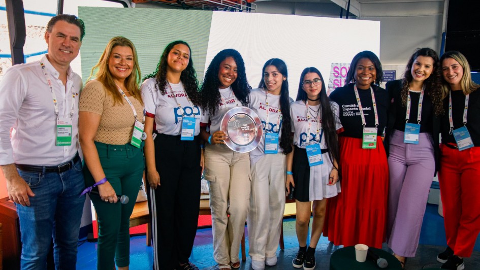 A imagem mostra um grupo de nove pessoas (sete mulheres e dois homens) posando para uma foto comemorativa em um ambiente de evento. No centro, um grupo de jovens mulheres segura um trof&eacute;u em formato de prato prateado, celebrando uma premia&ccedil;&atilde;o ou conquista.

Detalhes das Pessoas e Vestimentas
As Estudantes: No centro da foto, quatro jovens representam um grupo. Elas vestem camisetas brancas uniformizadas com logotipos azuis e vermelhos onde se l&ecirc; "POD" e refer&ecirc;ncias ao "Centro da Juventude Alvorada". Uma delas, no meio, segura orgulhosamente o pr&ecirc;mio prateado circular.

Autoridades e Organizadores: Ladeando as estudantes, est&atilde;o homens e mulheres em trajes casuais-sociais (camisas polo, blusas de tric&ocirc; e cal&ccedil;as coloridas em tons de verde, rosa e vermelho). Todos os presentes na imagem utilizam crach&aacute;s oficiais com cord&otilde;es coloridos pendurados no pesco&ccedil;o, identificando-os como participantes ou organizadores do evento.

Express&otilde;es: O clima &eacute; de celebra&ccedil;&atilde;o e alegria, com todos os integrantes sorrindo para a c&acirc;mera.

Ambiente e Fundo
Cen&aacute;rio: O grupo est&aacute; posicionado &agrave; frente de um grande painel de LED que exibe uma luz branca brilhante, criando um fundo limpo. No canto direito do painel, &eacute; poss&iacute;vel ver parte do logotipo do evento.

Localiza&ccedil;&atilde;o: O ambiente sugere ser um pavilh&atilde;o ou espa&ccedil;o de confer&ecirc;ncias moderno. No ch&atilde;o, em primeiro plano, v&ecirc;-se um microfone e um copo descart&aacute;vel sobre uma pequena mesa, indicando que uma apresenta&ccedil;&atilde;o ou cerim&ocirc;nia acabou de ocorrer.

Composi&ccedil;&atilde;o
A foto &eacute; frontal e enquadra o grupo da cintura para cima, com as pessoas levemente voltadas para o centro, onde est&aacute; o trof&eacute;u. A ilumina&ccedil;&atilde;o &eacute; clara e direta, destacando as cores das roupas e a textura do pr&ecirc;mio met&aacute;lico.