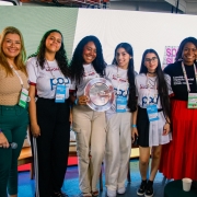 A imagem mostra um grupo de nove pessoas (sete mulheres e dois homens) posando para uma foto comemorativa em um ambiente de evento. No centro, um grupo de jovens mulheres segura um trof&eacute;u em formato de prato prateado, celebrando uma premia&ccedil;&atilde;o ou conquista.

Detalhes das Pessoas e Vestimentas
As Estudantes: No centro da foto, quatro jovens representam um grupo. Elas vestem camisetas brancas uniformizadas com logotipos azuis e vermelhos onde se l&ecirc; "POD" e refer&ecirc;ncias ao "Centro da Juventude Alvorada". Uma delas, no meio, segura orgulhosamente o pr&ecirc;mio prateado circular.

Autoridades e Organizadores: Ladeando as estudantes, est&atilde;o homens e mulheres em trajes casuais-sociais (camisas polo, blusas de tric&ocirc; e cal&ccedil;as coloridas em tons de verde, rosa e vermelho). Todos os presentes na imagem utilizam crach&aacute;s oficiais com cord&otilde;es coloridos pendurados no pesco&ccedil;o, identificando-os como participantes ou organizadores do evento.

Express&otilde;es: O clima &eacute; de celebra&ccedil;&atilde;o e alegria, com todos os integrantes sorrindo para a c&acirc;mera.

Ambiente e Fundo
Cen&aacute;rio: O grupo est&aacute; posicionado &agrave; frente de um grande painel de LED que exibe uma luz branca brilhante, criando um fundo limpo. No canto direito do painel, &eacute; poss&iacute;vel ver parte do logotipo do evento.

Localiza&ccedil;&atilde;o: O ambiente sugere ser um pavilh&atilde;o ou espa&ccedil;o de confer&ecirc;ncias moderno. No ch&atilde;o, em primeiro plano, v&ecirc;-se um microfone e um copo descart&aacute;vel sobre uma pequena mesa, indicando que uma apresenta&ccedil;&atilde;o ou cerim&ocirc;nia acabou de ocorrer.

Composi&ccedil;&atilde;o
A foto &eacute; frontal e enquadra o grupo da cintura para cima, com as pessoas levemente voltadas para o centro, onde est&aacute; o trof&eacute;u. A ilumina&ccedil;&atilde;o &eacute; clara e direta, destacando as cores das roupas e a textura do pr&ecirc;mio met&aacute;lico.