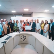 A imagem mostra uma reuni&atilde;o institucional em uma sala de reuni&otilde;es ampla, com mesa em formato semicircular. Um grupo numeroso est&aacute; reunido em p&eacute; atr&aacute;s da mesa, enquanto, ao centro, duas pessoas seguram um documento aberto.