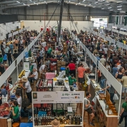 A imagem mostra uma feira agroindustrial realizada em um grande pavilh&atilde;o coberto, com corredores longos e estandes organizados em fileiras paralelas. Os estandes exibem produtos aliment&iacute;cios, bebidas, utens&iacute;lios e itens artesanais, com bancadas de madeira e sinaliza&ccedil;&atilde;o suspensa indicando marcas e expositores. O espa&ccedil;o est&aacute; intensamente ocupado por visitantes circulando pelos corredores. 