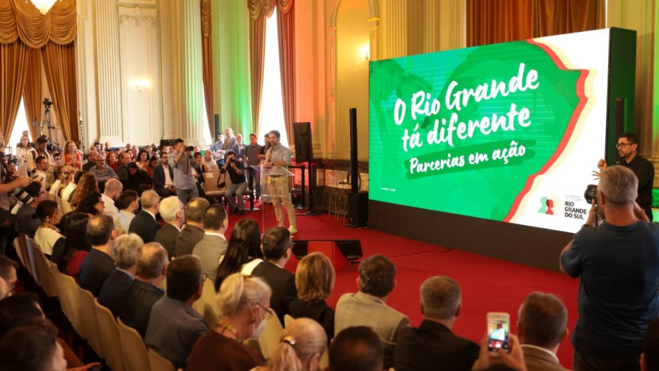A imagem mostra um evento institucional em um sal&atilde;o hist&oacute;rico, com plateia ocupando fileiras de cadeiras diante de um palco. &Agrave; direita, um tel&atilde;o grande exibe a mensagem &ldquo;O Rio Grande t&aacute; diferente &ndash; Parcerias em a&ccedil;&atilde;o&rdquo; sobre fundo verde com marca visual do governo estadual. &Agrave; esquerda do tel&atilde;o, o governador est&aacute; posicionado com microfone, participando da apresenta&ccedil;&atilde;o. 