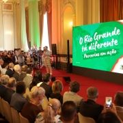 A imagem mostra um evento institucional em um sal&atilde;o hist&oacute;rico, com plateia ocupando fileiras de cadeiras diante de um palco. &Agrave; direita, um tel&atilde;o grande exibe a mensagem &ldquo;O Rio Grande t&aacute; diferente &ndash; Parcerias em a&ccedil;&atilde;o&rdquo; sobre fundo verde com marca visual do governo estadual. &Agrave; esquerda do tel&atilde;o, o governador est&aacute; posicionado com microfone, participando da apresenta&ccedil;&atilde;o. 