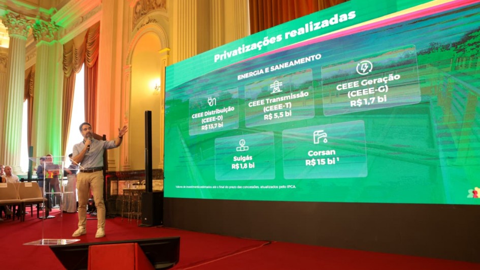 Esta &eacute; uma descri&ccedil;&atilde;o detalhada da imagem para fins de acessibilidade:

Vis&atilde;o Geral
A imagem mostra uma apresenta&ccedil;&atilde;o oficial realizada em um sal&atilde;o cl&aacute;ssico e imponente. &Agrave; esquerda, um homem jovem est&aacute; de p&eacute; em um palco, falando ao microfone e gesticulando em dire&ccedil;&atilde;o a um grande tel&atilde;o de LED posicionado &agrave; direita, que exibe dados sobre privatiza&ccedil;&otilde;es.

Detalhes do Palestrante e Palco
O Palestrante: O homem tem pele clara, cabelos e barba escuros. Ele veste uma camisa social azul clara com as mangas dobradas, cal&ccedil;as de sarja bege e t&ecirc;nis brancos. Ele segura o microfone com a m&atilde;o direita e estende o bra&ccedil;o esquerdo em dire&ccedil;&atilde;o &agrave; tela.

O Palco: O ch&atilde;o do palco &eacute; coberto por um carpete vermelho vibrante. &Agrave; frente do palestrante, h&aacute; um p&uacute;lpito de acr&iacute;lico transparente e, no ch&atilde;o, um monitor de retorno preto voltado para ele.

Conte&uacute;do do Tel&atilde;o
O tel&atilde;o apresenta um fundo verde brilhante com o t&iacute;tulo "Privatiza&ccedil;&otilde;es realizadas" no topo. Abaixo, na se&ccedil;&atilde;o "ENERGIA E SANEAMENTO", cinco blocos informativos listam empresas e valores em bilh&otilde;es de reais:

CEEE Distribui&ccedil;&atilde;o (CEEE-D): R$ 13,7 bi

CEEE Transmiss&atilde;o (CEEE-T): R$ 5,5 bi

CEEE Gera&ccedil;&atilde;o (CEEE-G): R$ 1,7 bi

Sulg&aacute;s: R$ 1,8 bi

Corsan: R$ 15 bi

Ambiente e Arquitetura
O local possui uma arquitetura neocl&aacute;ssica luxuosa, com colunas altas de capit&eacute;is trabalhados, paredes decoradas com molduras douradas e pinturas em relevo (afrescos). Grandes janelas com cortinas pesadas em tons de dourado e marrom comp&otilde;em o fundo. A ilumina&ccedil;&atilde;o do sal&atilde;o &eacute; quente, contrastando com a luz verde intensa emitida pelo tel&atilde;o.