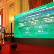 Esta &eacute; uma descri&ccedil;&atilde;o detalhada da imagem para fins de acessibilidade:

Vis&atilde;o Geral
A imagem mostra uma apresenta&ccedil;&atilde;o oficial realizada em um sal&atilde;o cl&aacute;ssico e imponente. &Agrave; esquerda, um homem jovem est&aacute; de p&eacute; em um palco, falando ao microfone e gesticulando em dire&ccedil;&atilde;o a um grande tel&atilde;o de LED posicionado &agrave; direita, que exibe dados sobre privatiza&ccedil;&otilde;es.

Detalhes do Palestrante e Palco
O Palestrante: O homem tem pele clara, cabelos e barba escuros. Ele veste uma camisa social azul clara com as mangas dobradas, cal&ccedil;as de sarja bege e t&ecirc;nis brancos. Ele segura o microfone com a m&atilde;o direita e estende o bra&ccedil;o esquerdo em dire&ccedil;&atilde;o &agrave; tela.

O Palco: O ch&atilde;o do palco &eacute; coberto por um carpete vermelho vibrante. &Agrave; frente do palestrante, h&aacute; um p&uacute;lpito de acr&iacute;lico transparente e, no ch&atilde;o, um monitor de retorno preto voltado para ele.

Conte&uacute;do do Tel&atilde;o
O tel&atilde;o apresenta um fundo verde brilhante com o t&iacute;tulo "Privatiza&ccedil;&otilde;es realizadas" no topo. Abaixo, na se&ccedil;&atilde;o "ENERGIA E SANEAMENTO", cinco blocos informativos listam empresas e valores em bilh&otilde;es de reais:

CEEE Distribui&ccedil;&atilde;o (CEEE-D): R$ 13,7 bi

CEEE Transmiss&atilde;o (CEEE-T): R$ 5,5 bi

CEEE Gera&ccedil;&atilde;o (CEEE-G): R$ 1,7 bi

Sulg&aacute;s: R$ 1,8 bi

Corsan: R$ 15 bi

Ambiente e Arquitetura
O local possui uma arquitetura neocl&aacute;ssica luxuosa, com colunas altas de capit&eacute;is trabalhados, paredes decoradas com molduras douradas e pinturas em relevo (afrescos). Grandes janelas com cortinas pesadas em tons de dourado e marrom comp&otilde;em o fundo. A ilumina&ccedil;&atilde;o do sal&atilde;o &eacute; quente, contrastando com a luz verde intensa emitida pelo tel&atilde;o.