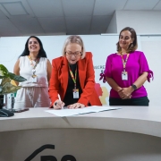 A imagem mostra a secret&aacute;ria da Sa&uacute;de assinando um documento sobre uma mesa clara em ambiente institucional, enquanto duas outras pessoas permanecem ao fundo observando.