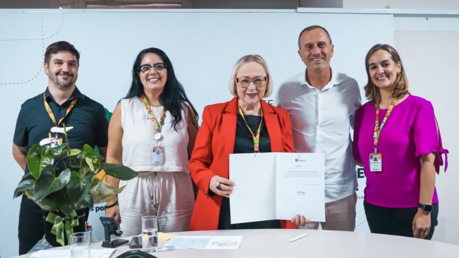 A imagem mostra um grupo de cinco pessoas reunido atr&aacute;s de uma mesa semicircular em ambiente institucional. A pessoa ao centro segura um documento aberto, exibido voltado para a c&acirc;mera.